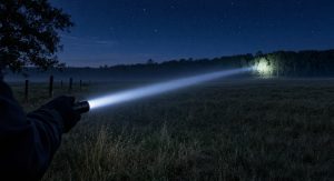 Long range throw flashlight beam reaching across a dark field to a distant tree line